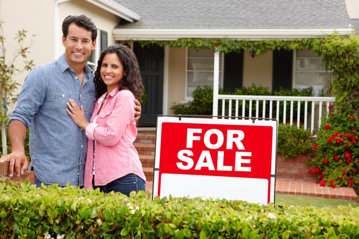 Couple standing outside of their home with for sale sign